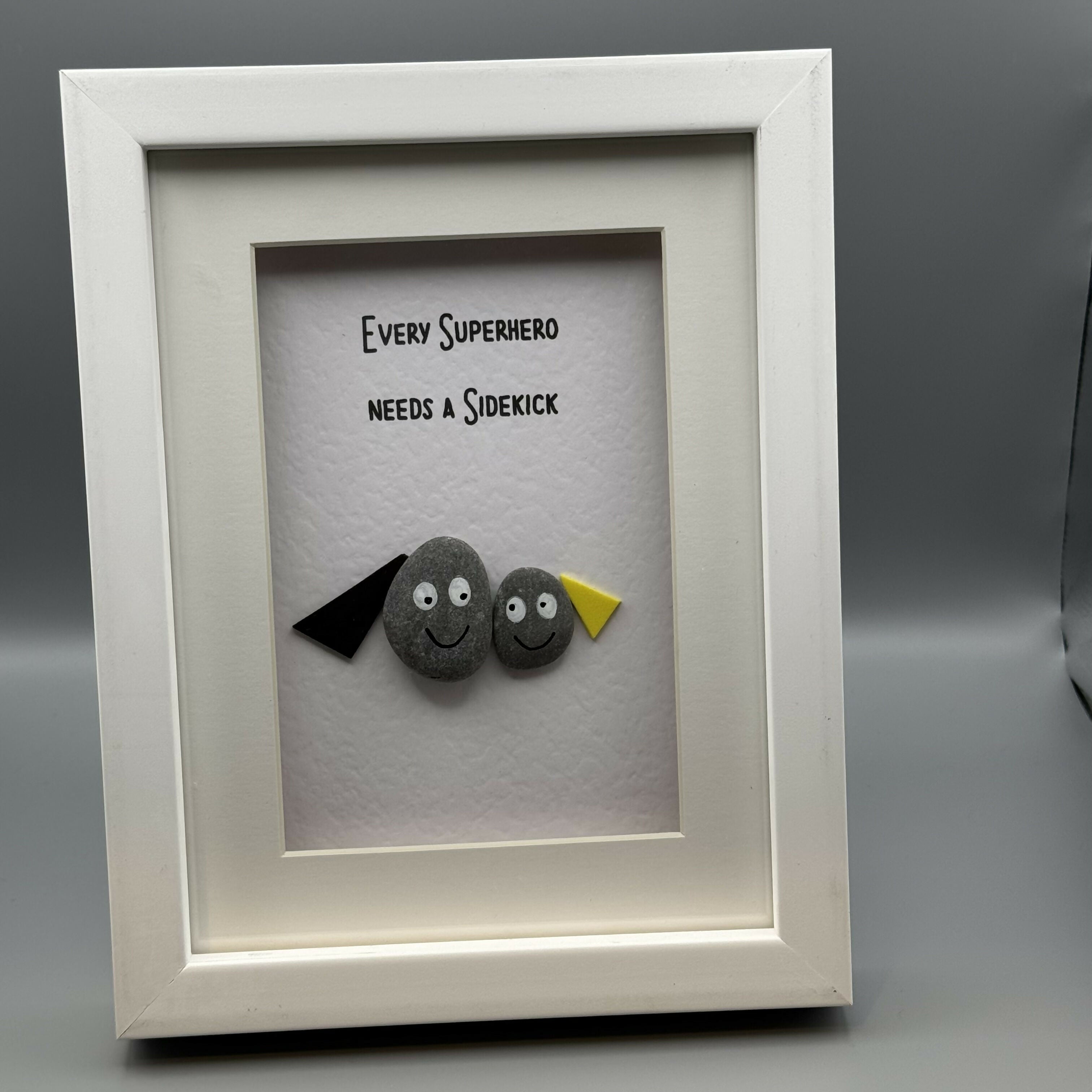 White frame with two pebbles with faces and capes, text says 'every superhero needs a sidekick'.
