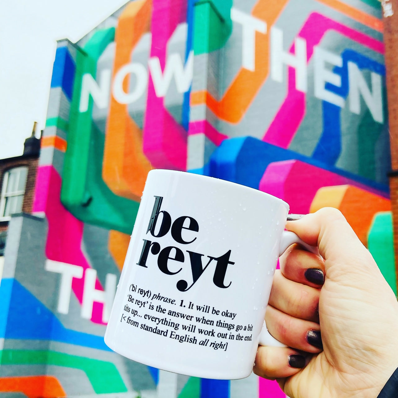 A 'be reyt' mug in front of colourful building.