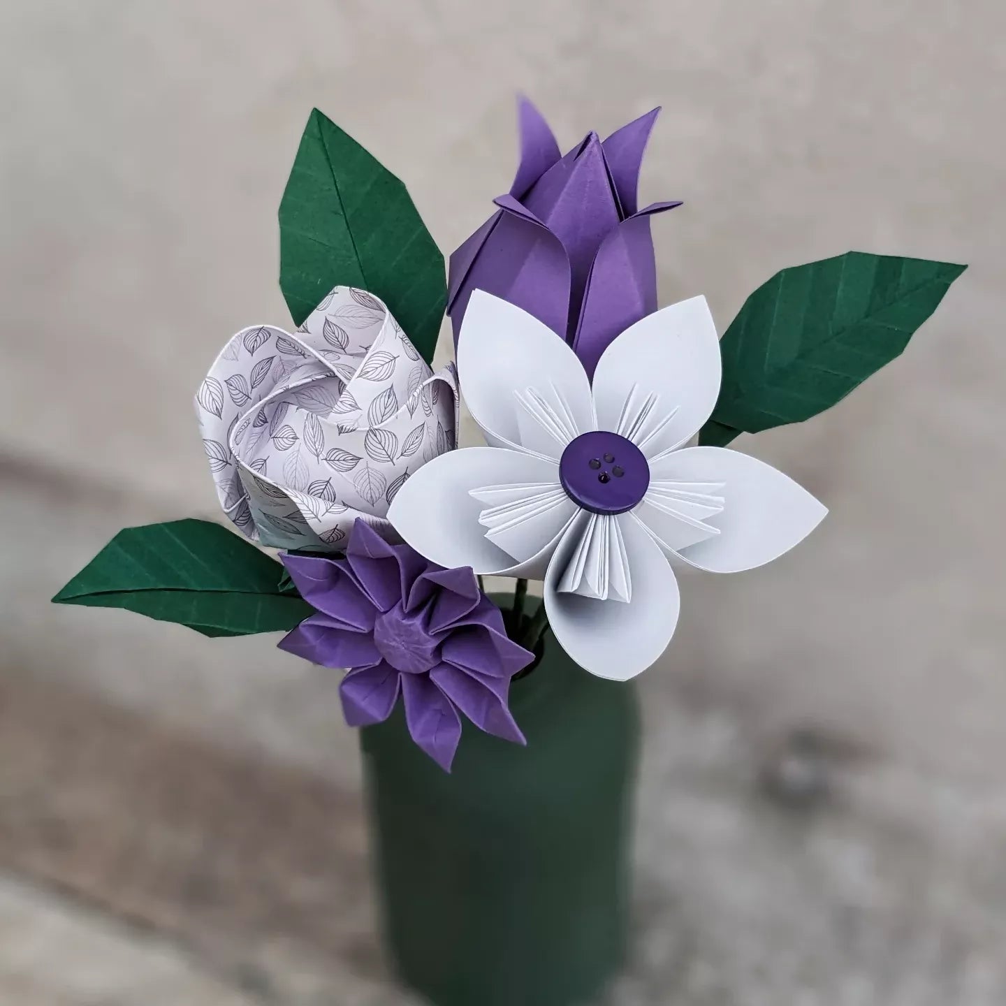 bouquet of paper flowers featuring a white daisy, purple tulip, purple gerbera, white patterned rose and three dark green folded origami leaves